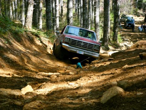2005 Jeep Wrangler - A red Ford pickup truck is navigating a steep, muddy, and rocky off-road trail through a forest. The truck is angled sharply, with its fron…