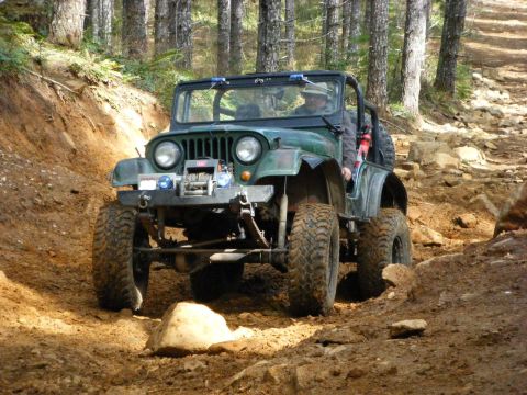 2005 Jeep Wrangler - A green Jeep Wrangler is driving up a steep, rocky, and muddy trail in a forest. The Jeep has large off-road tires, a winch on the front bu…