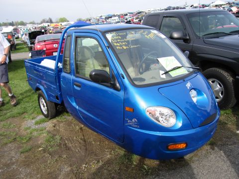 2001 Ford Crown Victoria - A blue three-wheeled microcar is parked at an outdoor event with other vehicles and tents in the background.