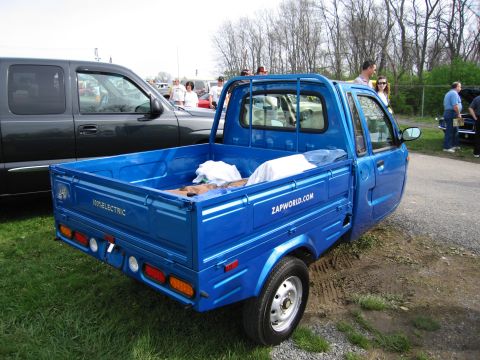 2001 Ford Crown Victoria - A bright blue three-wheeled microtruck is parked on grass next to a dark pickup truck.