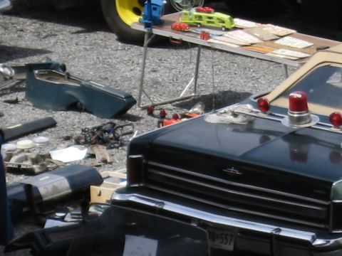 2001 Ford Crown Victoria - A dark sedan with emergency lights is parked on a gravel lot, surrounded by various car parts and items on a table.