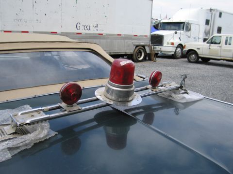 2001 Ford Crown Victoria - Emergency lights are mounted on the hood of a dark sedan, with trucks in the background.