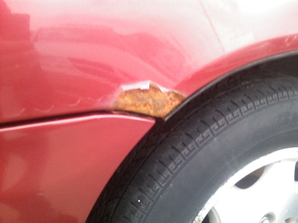 Rust hole bubbling through red car body panel above rear wheel arch