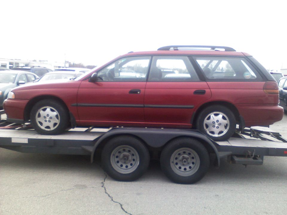 Maroon Subaru Legacy wagon loaded on a tandem-axle open car trailer, side view