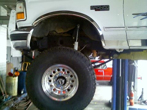 1996 Ford Bronco - A white 1996 Ford Bronco is shown on a lift, highlighting its lifted suspension and large tire. Another vehicle is visible in the backgroun…