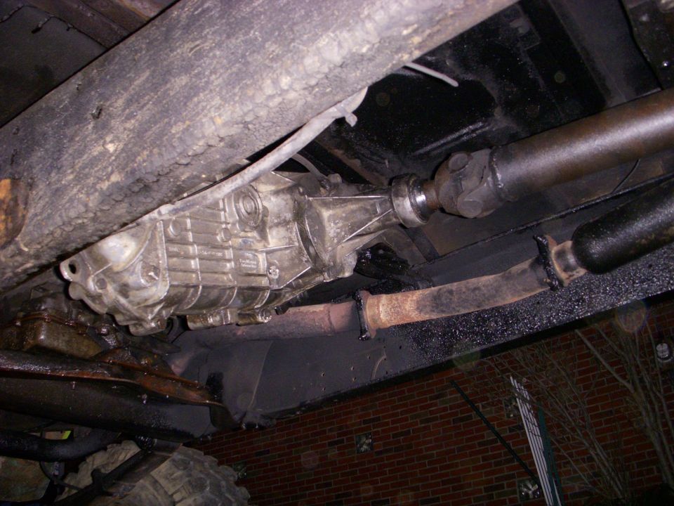 Underside view of a vehicle's transmission and driveshaft. Rusty exhaust pipe visible