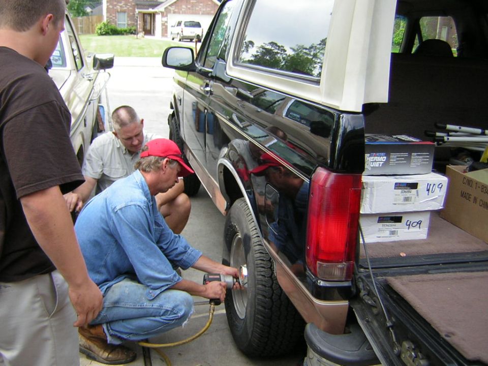 Man with red hat uses impact wrench on black Ford Bronco wheel, another man watches, third man stands nearby