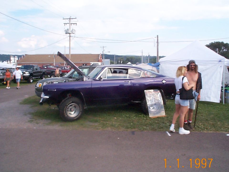 Purple fastback muscle car with open hood at an outdoor swap meet, people standing beside it