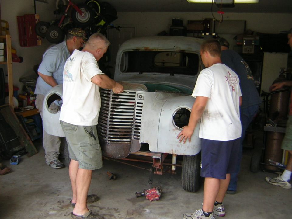 People working on a vintage truck's chrome grille and front end in a garage