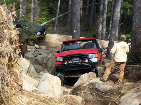 1989 Ford Bronco - Red 1989 Ford Bronco tackling a challenging rocky trail with a black SUV in the background.