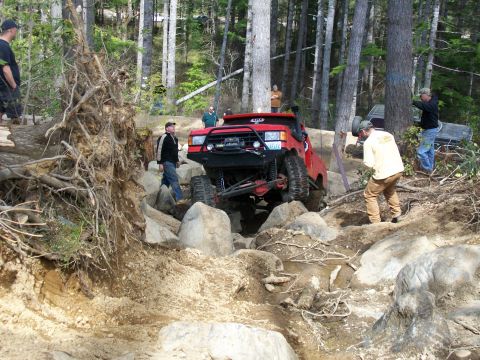 1989 Ford Bronco - Modified red SUV tackles a challenging rocky terrain with people observing.