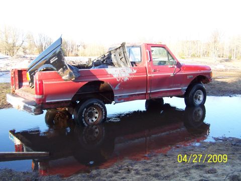 1989 Ford F-150 - A red 1989 Ford F-150 sits in a puddle, showing significant damage to its bed and body.