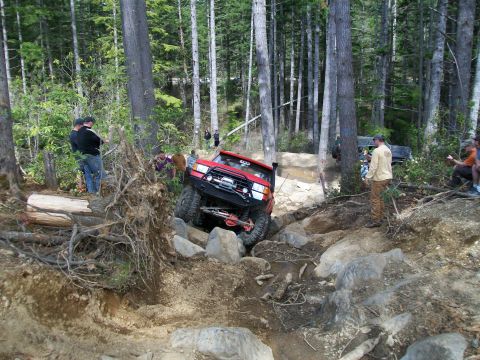 1989 Ford Bronco - A red off-road SUV tackles a challenging rocky terrain in a wooded area, with spectators watching.