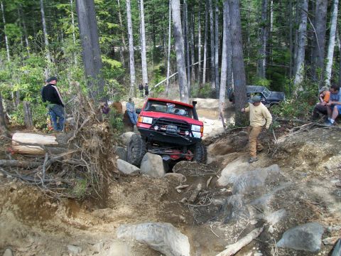 1989 Ford Bronco - A red 1989 Ford Bronco tackles a challenging rocky obstacle on an off-road trail, with spectators watching.