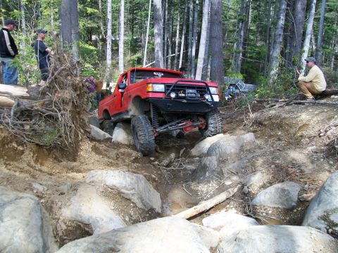 1989 Ford Bronco - A red pickup truck is shown navigating a challenging rocky terrain in a wooded area with spectators watching.
