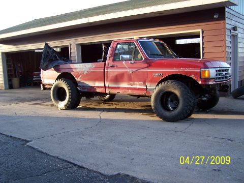 1989 Ford F-150 - This modified red Ford pickup truck, likely from the late 1980s, sits in front of a garage with its large tires visible.