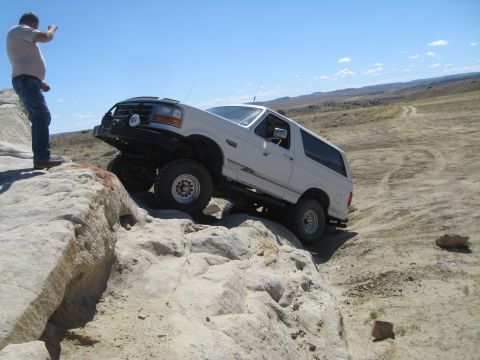 1993 Ford Bronco - White 1993 Ford Bronco navigates a rocky off-road obstacle under a clear blue sky.