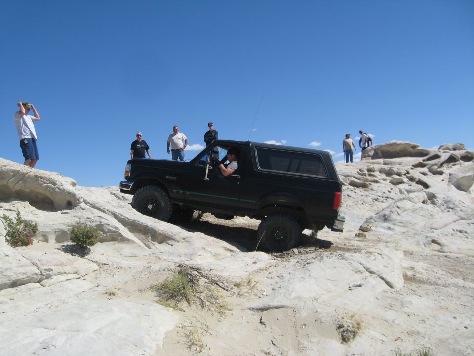Black 1993 Ford Bronco driving off-road on a bright, sunny day with several people watching