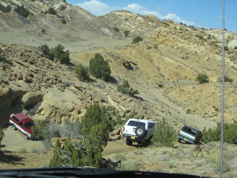 1993 Ford Bronco - Multiple SUVs navigate a challenging off-road trail through a , arid terrain under a partly cloudy sky.