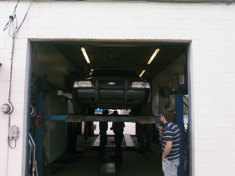 Dark Ford Crown Victoria raised on a lift in a garage bay, seen head-on with people standing below