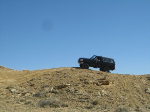 1993 Ford Bronco - A black Ford Bronco is driving up a steep, dusty hill under a clear blue sky. The vehicle is angled upwards, showcasing its off-road capabi…