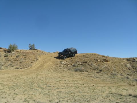 1993 Ford Bronco - A black 1993 Ford Bronco is driving up a dirt hill on a sunny day. The vehicle is viewed from the front-right angle, with a clear blue sky…