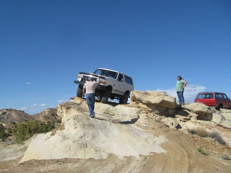 White Ford Bronco on a rocky hill with a person, red SUV in background.