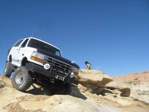 1993 Ford Bronco - A white 1993 Ford Bronco is shown off-roading on a rocky incline under a clear blue sky. A person in a green and white patterned shirt is v…