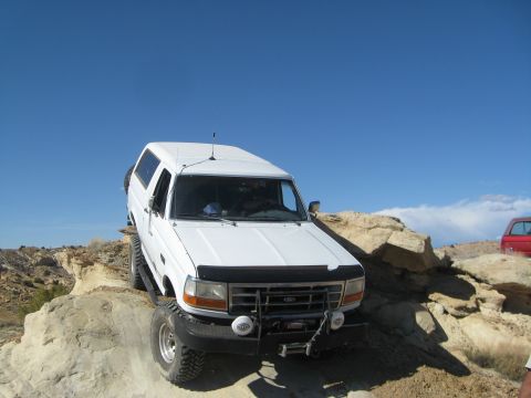 1993 Ford Bronco - A white Ford Bronco is navigating a rocky, off-road trail under a clear blue sky. The vehicle is angled on a rock, with its front tires vis…