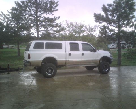 2000 Ford F-250 - This lifted pickup truck features a crew cab and a white camper shell, parked on a wet surface.