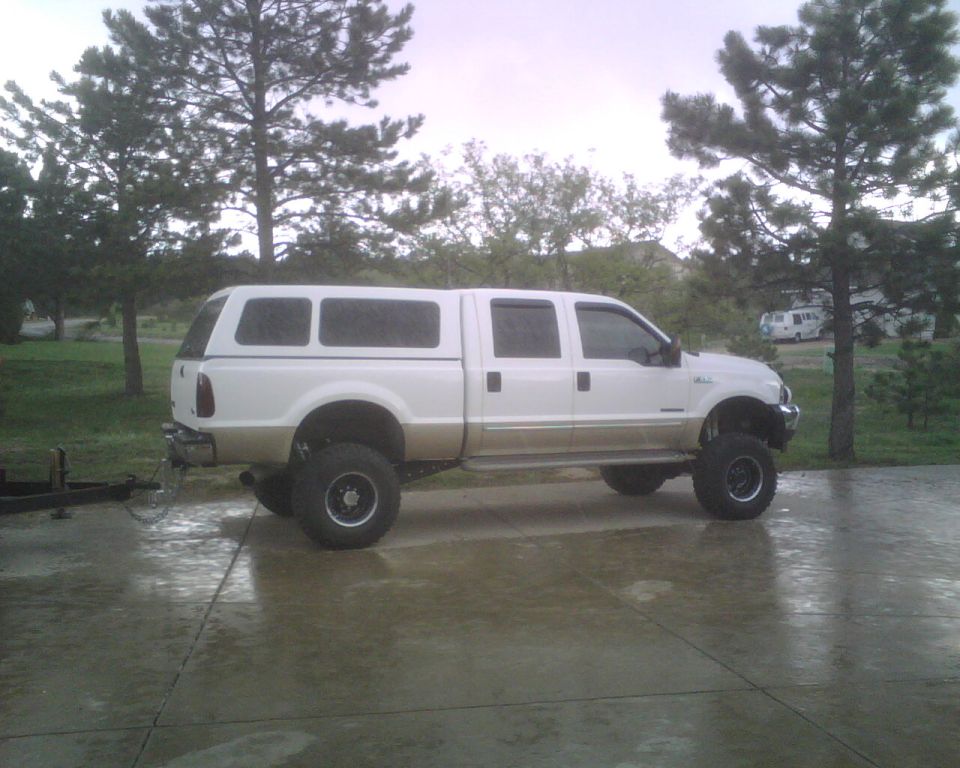 Lifted white and tan crew cab pickup truck with a white camper shell, parked on a wet driveway