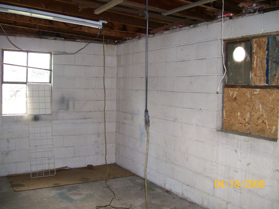 Empty whitewashed block garage corner with extension cord, boarded window and concrete floor