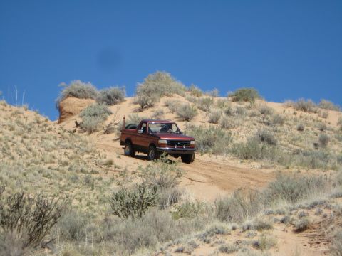 1993 Ford Bronco - Red 1993 Ford Bronco navigates a sandy trail in a desert environment under a bright blue sky.