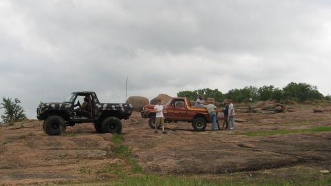 1979 Ford Bronco - Two off-road trucks are parked on a rocky terrain with a group of people gathered around the orange truck.