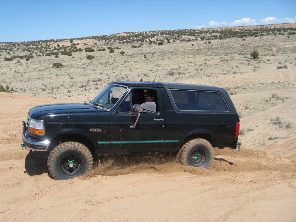 Black 1993 Ford Bronco driving off-road in sandy terrain, kicking up dust