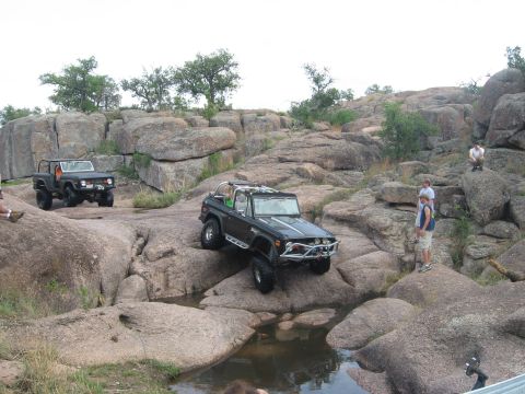 1979 Ford Bronco - Two Ford Broncos tackle a challenging off-road course with large rock formations and water.