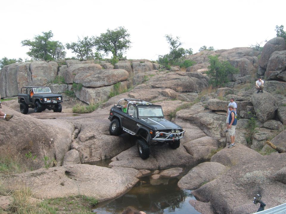 Two black Ford Broncos navigating rocky terrain with water obstacles. One Bronco is in the foreground, angled towards the viewer