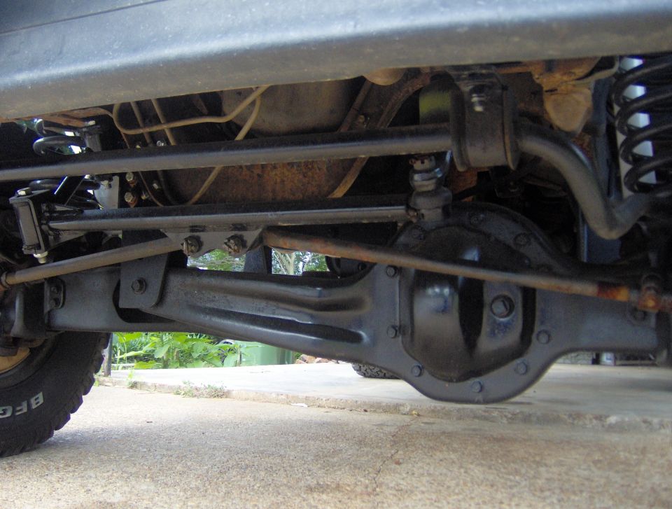 Underside of a vehicle showing the front axle, steering components, and suspension. Tire visible on the left
