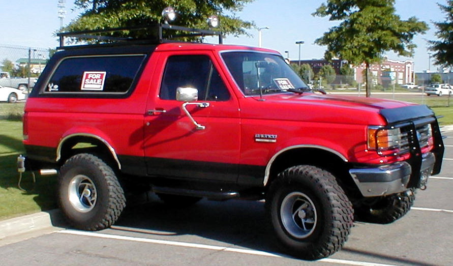 Red Ford Bronco SUV with roof rack, brush guard and For Sale signs, parked in a lot