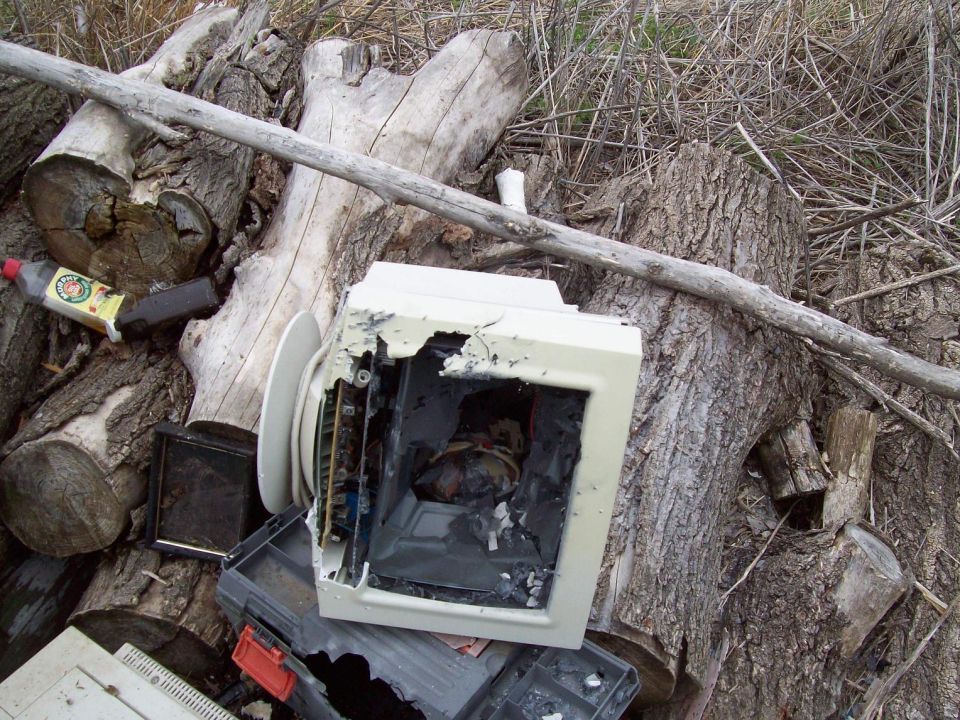 Broken computer monitor and other junk piled up against logs