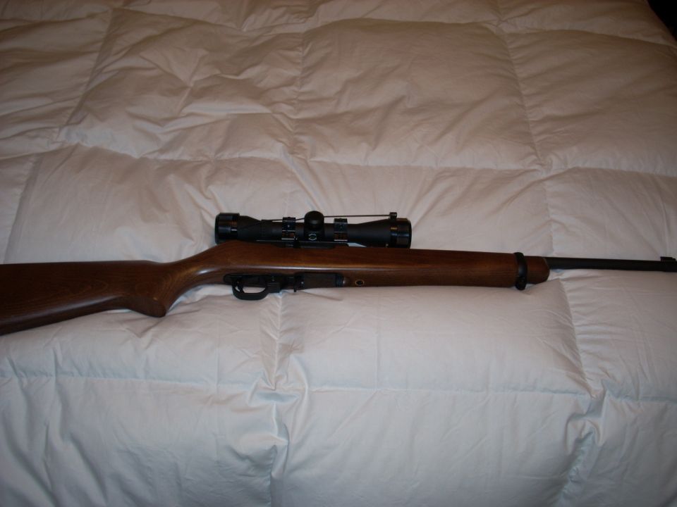 Wood-stocked rifle with black scope lying on a white quilted bedspread