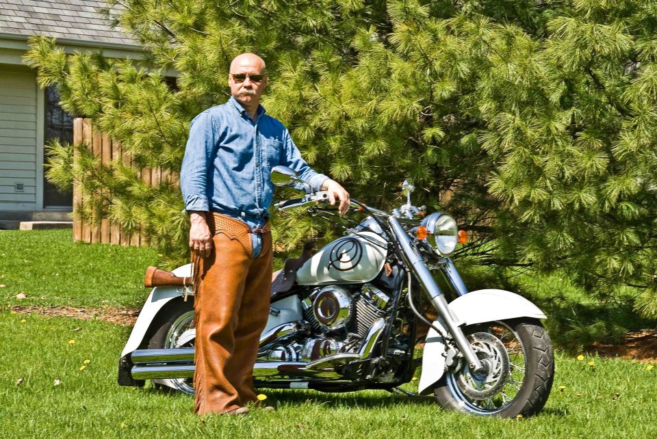 Man in denim shirt and chaps standing next to a 2006 Yamaha V Star Classic motorcycle on grass