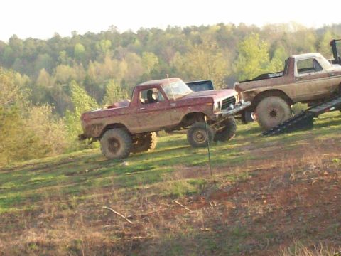 1978 Ford Bronco - Two lifted trucks are parked on a grassy, muddy incline with trees in the background.