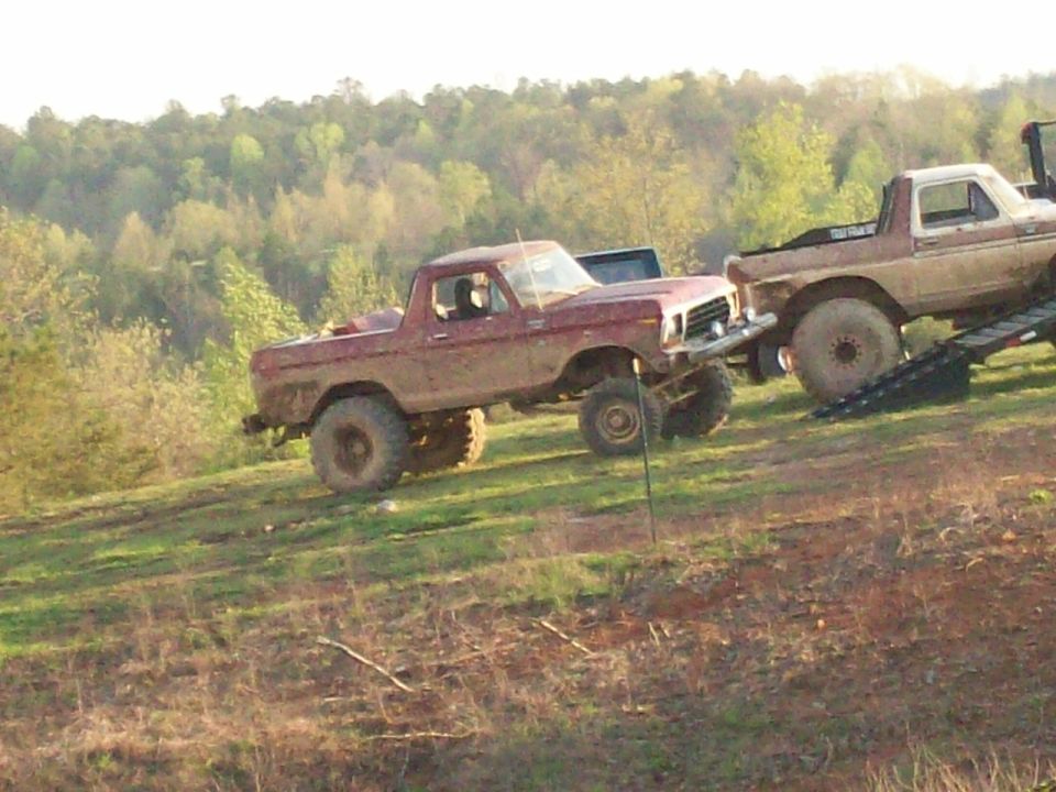 Muddy red Ford Bronco parked on a hill next to another lifted truck on a ramp