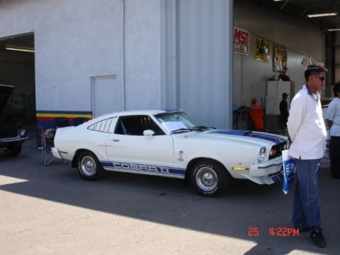1978 Ford Mustang II - A white 1978 Ford Mustang II Cobra with blue racing stripes is parked in front of a garage with a man standing nearby.
