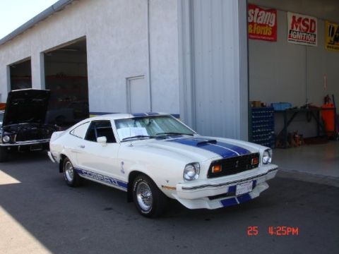 1978 Ford Mustang II - A white 1978 Ford Mustang II coupe with blue racing stripes is parked in front of a garage, with another car visible in the background.