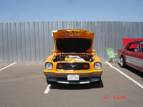 1978 Ford Mustang II - An orange coupe is parked in a lot with its hood up, next to a red coupe. A green chair sits between them.
