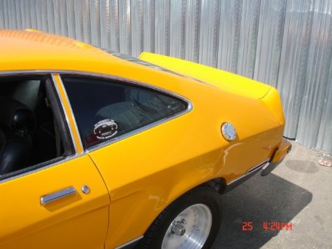 1978 Ford Mustang II - A yellow coupe is parked next to a corrugated metal wall, showing its rear quarter panel and side window.