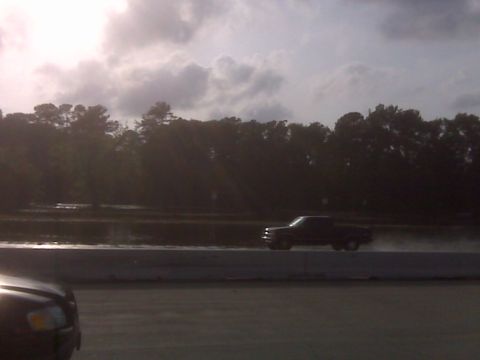 2008 Jeep Wrangler - A dark pickup truck is seen driving on a wet surface with trees lining the horizon under a cloudy sky.