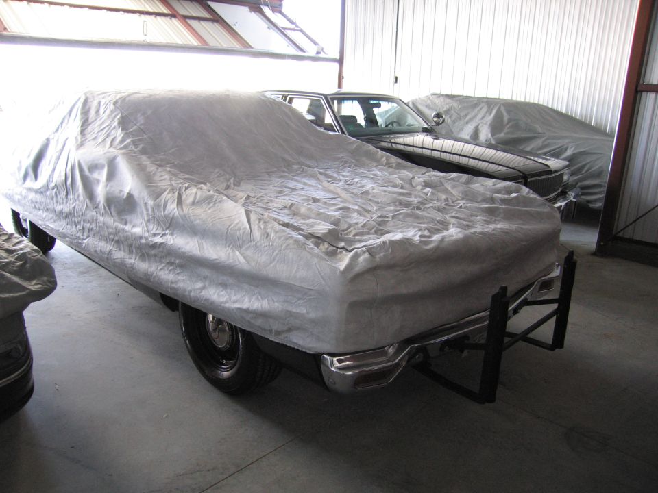 Several cars covered in tarps inside a garage. One car in the foreground has a chrome bumper and a black frame attached to the rear